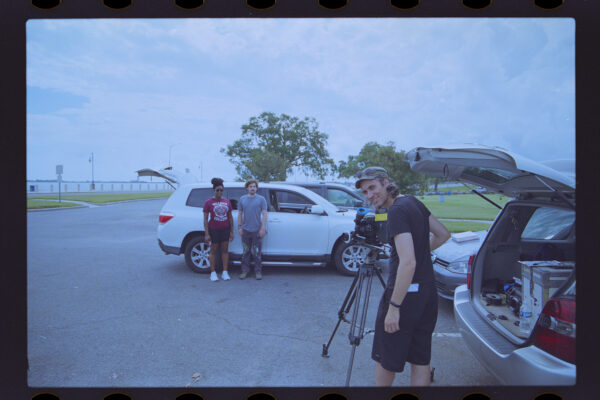 Ted turns around and smiles from camera while setting up a shot in a parking lot; April Louise and Casey Shaw in the background