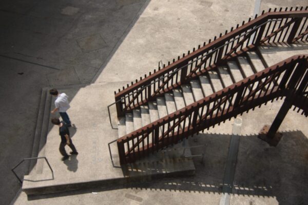 High angle geometric shot of Gold Connections descending outdoor staircase, walking toward sharp line of shadow