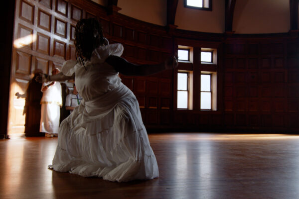 Woman in white dress dancing in open, wood-paneled circular room with light streaming through windows