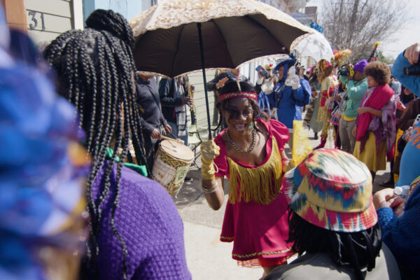 Woman in red and gold dress dancing with umbrella in a crowd on a sidewalk with drummers in the background