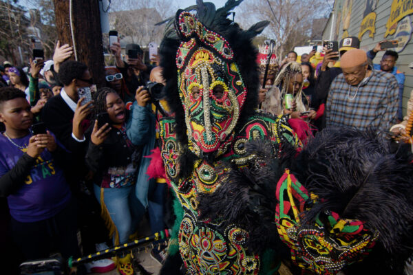 Man dressed in large, colorful beaded mask and outfit with black feathers looks into camera as excited crowd watches